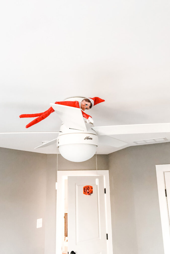 This image shows an elf on the shelf doll on a ceiling fan. This is one of the many elf on the shelf ideas for home included in this post.