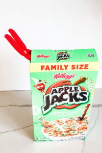 This image shows an elf on the shelf’s legs sticking out of a cereal box of Apple Jacks. This is one of the many elf on the shelf ideas for home included in this post.