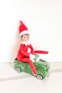 This image shows an elf on the elf doll sitting on a green truck Christmas decoration. This is one of the many elf on the shelf ideas for home included in this post.