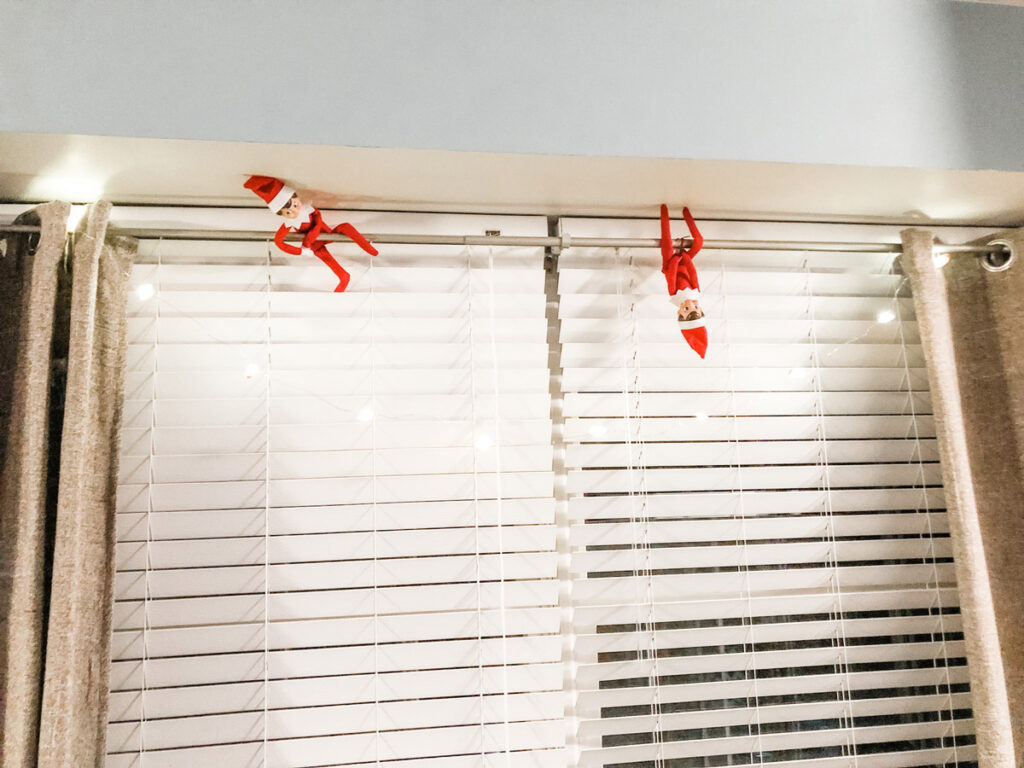 This image shows two elf on a shelf dolls hanging upside down off of a curtain rod with some Christmas lights strung across. This is one of the many elf on the shelf ideas for home included in this post.