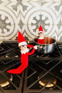 In this image, an elf on the shelf doll is pretending to cook another elf on the shelf doll in a pot. This is one of the many elf on the shelf ideas for home included in this post.