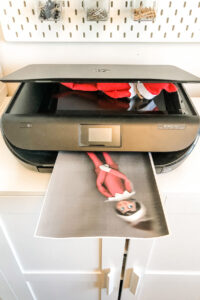 This image shows an elf on a shelf on a copier screen with the lid closed. In the printer tray there is a copy of the elf on a shelf printed on the paper. This is one of the many elf on the shelf ideas for home included in this post.