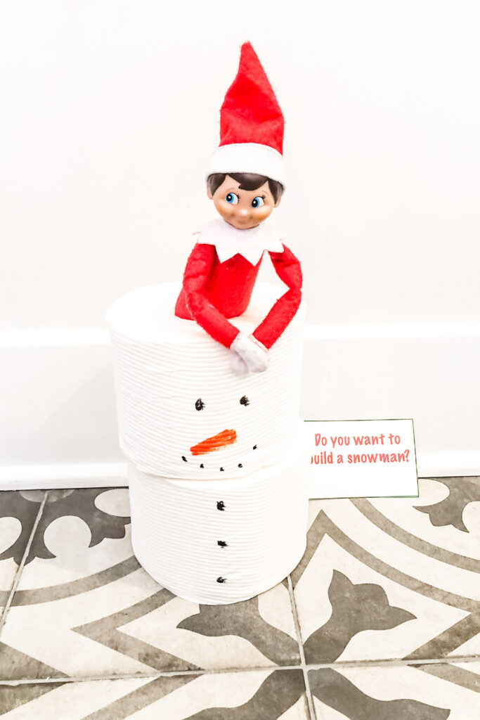 This image shows an elf stuck into 2 stacked rolls of toilet paper. They both having some drawings on them to look like some eyes, a carrot nose, smile, and buttons. There is a note next to the toilet paper rolls that says Do You want to build a snowman? This is one of the many elf on the shelf ideas for home included in this post.