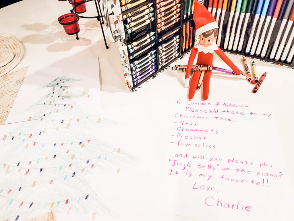 In this image there is an elf on the shelf doll pictured with a Christmas tree drawing. He is sitting on a note where he is asking his child he watches over to draw various elements on the tree. This is one of the many elf on the shelf ideas for home included in this post.