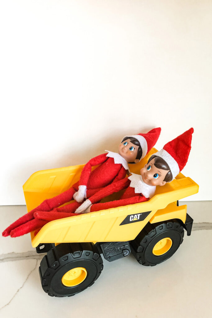 This image shows two elf on the shelf dolls inside of the back of a dump truck toy. This is one of the many elf on the shelf ideas for home included in this post.
