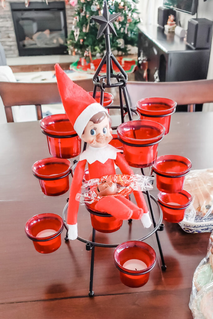 This image shows an elf on a shelf doll sitting in a candle holder covered in chocolate with a candy wrapper and a partially eaten piece of chocolate in his lap. This is one of the many elf on the shelf ideas for home included in this post.