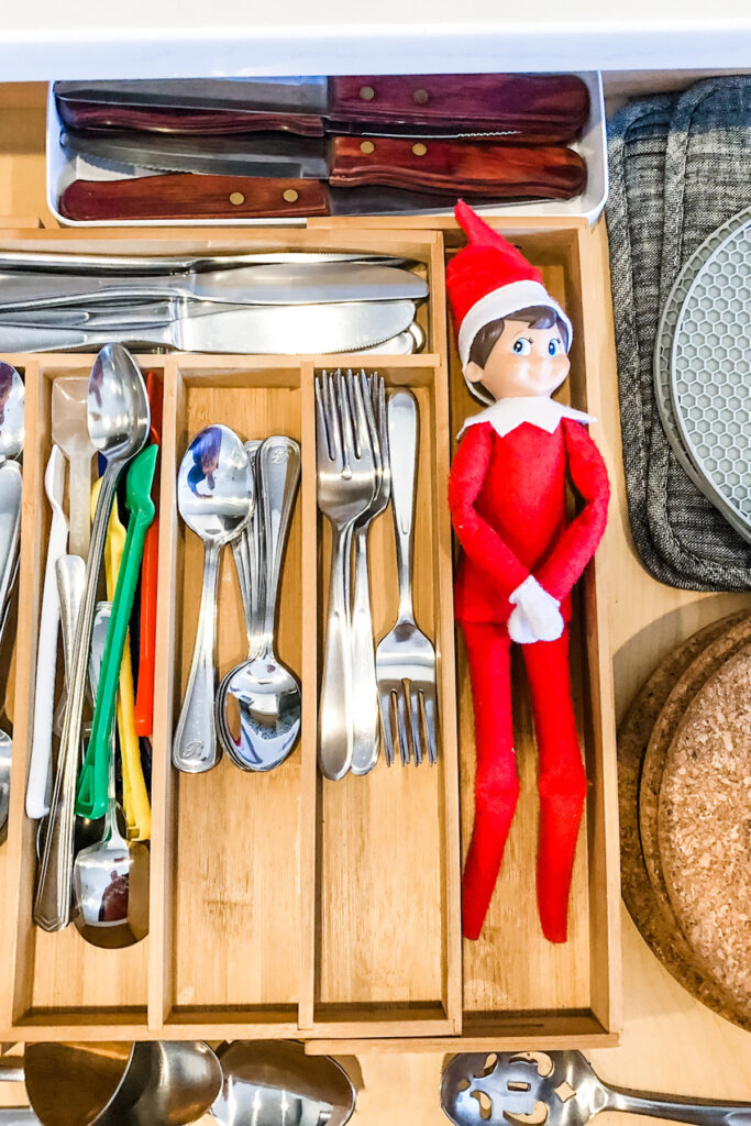 This image shows an elf on the shelf stuck inside of a utensil drawer. This is one of the many elf on the shelf ideas for home included in this post.