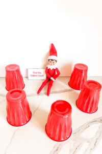 This image shows an elf sitting with 5 red cups surrounding him. He has a note that says find the hidden treat! This is one of the many elf on the shelf ideas for home included in this post.