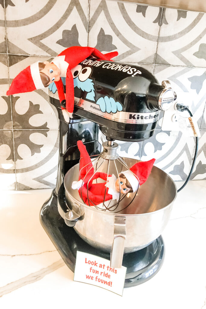 This image shows an elf inside of the whisk attachment on a kitchenmaid mixer. There is a second elf that appears to be pretending to turn on the mixer. There is a note that says look at this fun ride we found! This is one of the many elf on the shelf ideas for home included in this post.