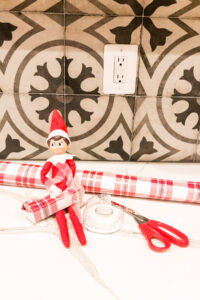 This image shows an elf on a shelf doll wrapping up a mini box in Christmas wrapping paper. This is one of the many elf on the shelf ideas for home included in this post.