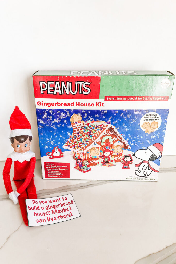 This image shows an elf sitting with a gingerbread house kit. There is a note that reads Do you want to build a gingerbread house? Maybe I can live there! This is one of the many elf on the shelf ideas for home included in this post.