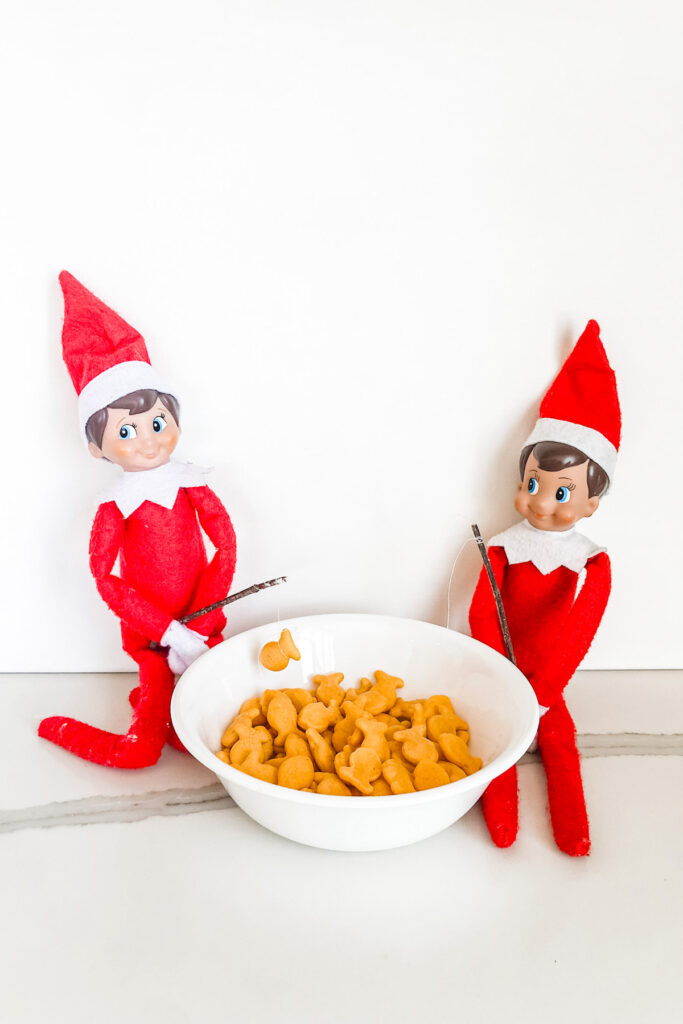 This image shows two elf on a shelf dolls fishing in goldfish. They are holding tiny twigs with thread attached. The one elf has a gold fish at the end of his string. This is one of the many elf on the shelf ideas for home included in this post.