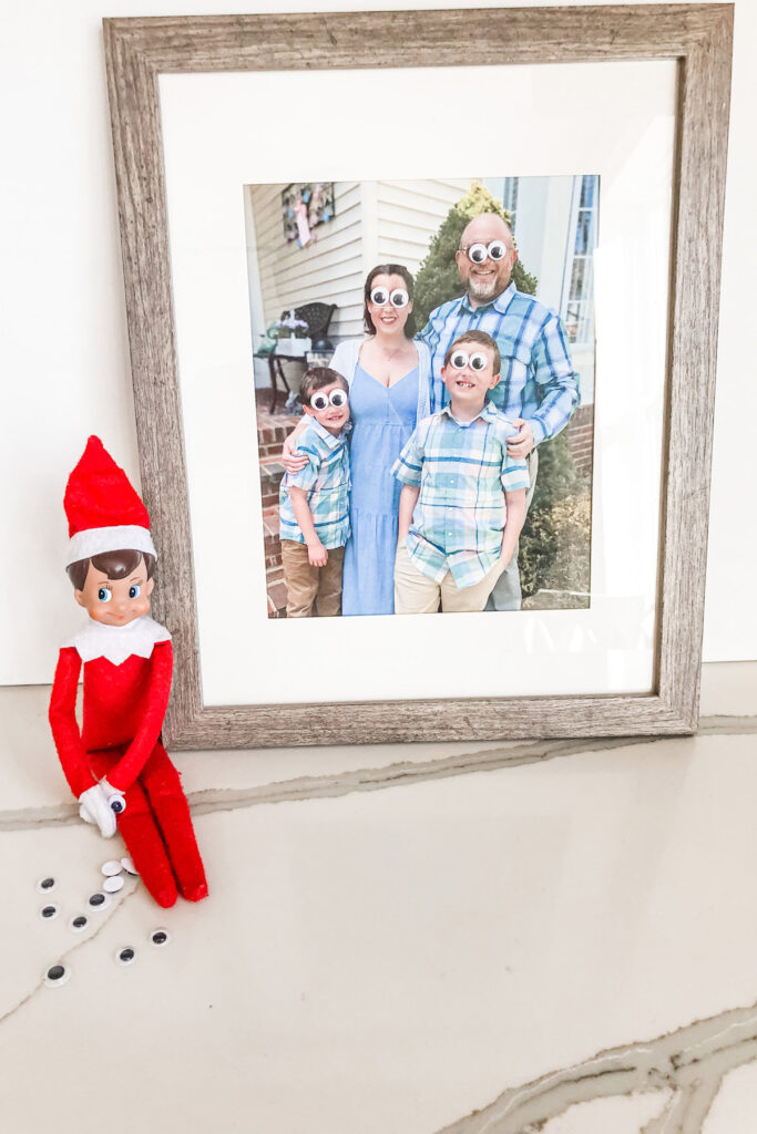 This image shows a family photo with googly eyes placed on where each person’s eyes are located. The elf on a shelf is sitting next to the picture with some googly eyes spread around. This is one of the many elf on the shelf ideas for home included in this post.