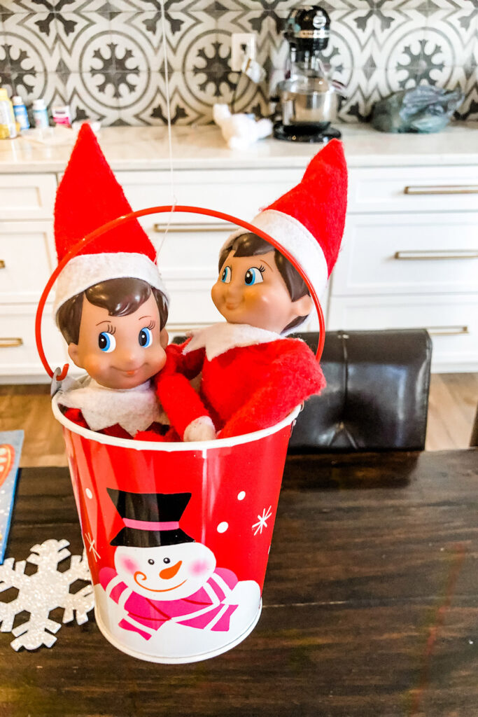 This image shows 2 elves inside of a tin basket hanging by string from a light fixture. This is one of the many elf on the shelf ideas for home included in this post.