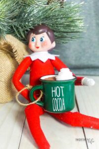 This image shoes an elf on the shelf doll drinking out of a fake miniature hot cocoa. This is one of the many elf on the shelf ideas for home included in this post.