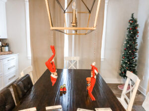 This image shows two elves hanging from a light fixture. This is one of the many elf on the shelf ideas for home included in this post.