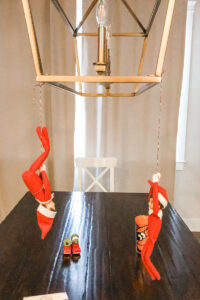 This image shows two elves hanging from a light fixture. This is one of the many elf on the shelf ideas for home included in this post.