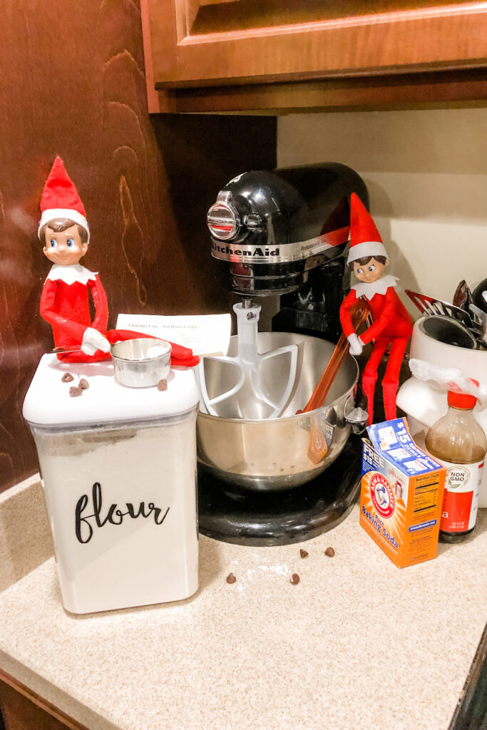 This image shows 2 elves pretending to bake cookies. There is a canister of flour out with the word flour on it. This is one of the many elf on the shelf ideas for home included in this post.