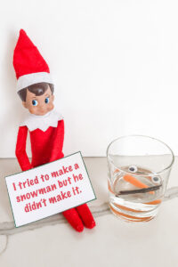 This image shows an elf on a shelf with a note that says I tried to make a snowman but he didn’t make it. Next to him is a glass of water with two twigs, 2 googly eyes, and 1 baby carrot. This is one of the many elf on the shelf ideas for home included in this post.
