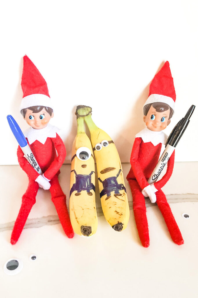 This image shows 2 elf on a shelf dolls holding Sharpe markers. Inbetween them are 2 bananas that have been drawn on to look like minions. This is one of the many elf on the shelf ideas for home included in this post.