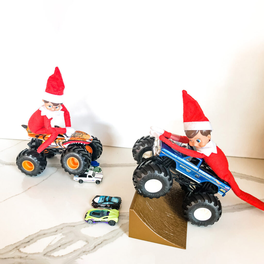 This image shows 2 elf on a shelf dolls riding toy monster trucks driving over top of cars. This is one of the many elf on the shelf ideas for home included in this post.