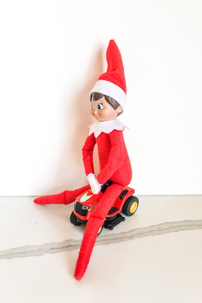 This image shows an elf on the shelf doll riding on a toy mower. This is one of the many elf on the shelf ideas for home included in this post.