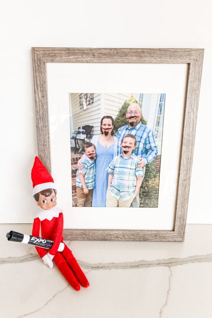 This image shows a family picture with an elf on the shelf sitting next to it holding a dry erase marker. The family picture has mustaches and beards drawn on each person. This is one of the many elf on the shelf ideas for home included in this post.