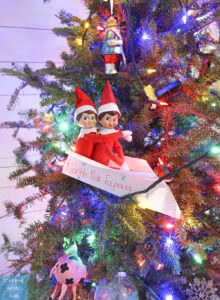 This image shows 2 elf on the shelf dolls on a paper airplane that has landed in a Christmas tree. This is one of the many elf on the shelf ideas for home included in this post.