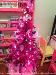This image is of a pink Christmas tree with a tiara at the top with an elf on the shelf doll holding onto the tiara. This is one of the many elf on the shelf ideas for home included in this post.
