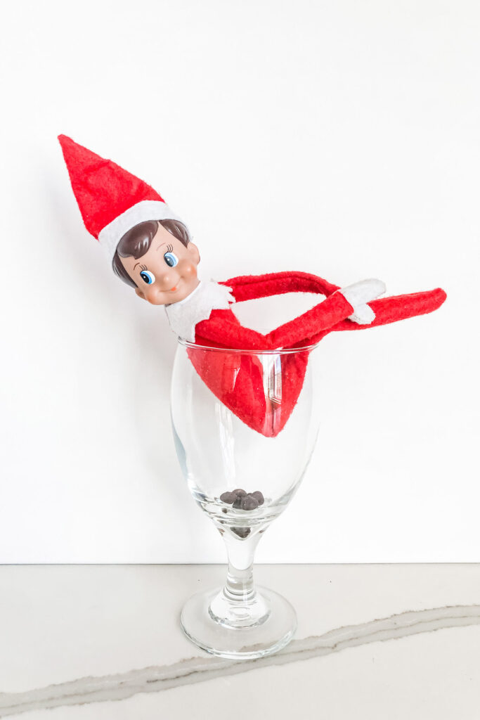 This image shows an elf on the shelf sitting in a glass appearing to be pooping, but it is in reality it is chocolate chips. This is one of the many elf on the shelf ideas for home included in this post