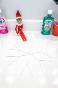 This image shows an elf on the shelf doll that has created a snowflake out of qtips This is one of the many elf on the shelf ideas for home included in this post.