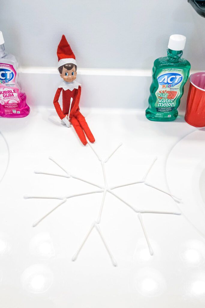 This image shows an elf on the shelf doll that has created a snowflake out of qtips This is one of the many elf on the shelf ideas for home included in this post.