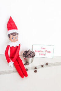 This image shows an elf on the shelf sitting next to a little silver container of chocolate chips. Next to that is a sign that says free reindeer poop! Enjoy! This is one of the many elf on the shelf ideas for home included in this post.