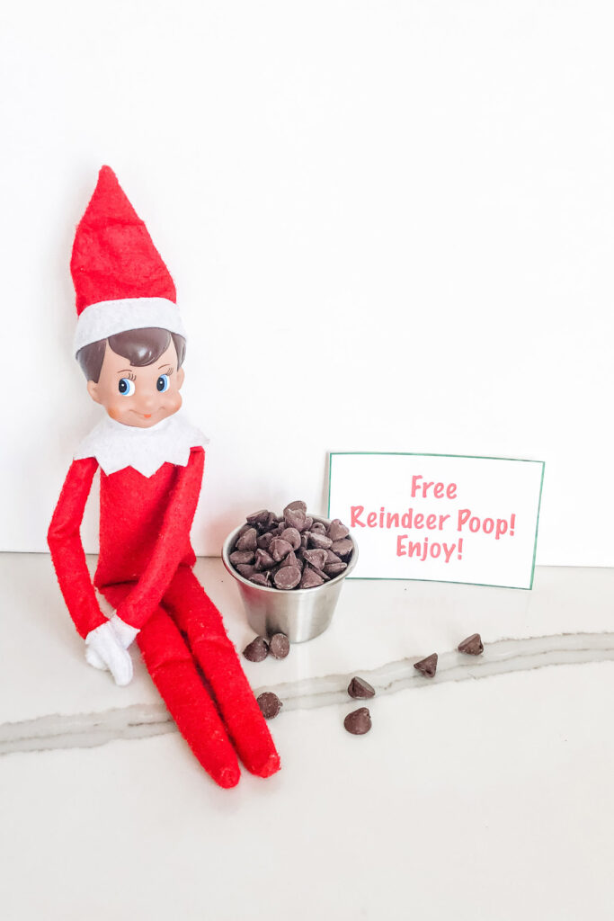 This image shows an elf on the shelf sitting next to a little silver container of chocolate chips. Next to that is a sign that says free reindeer poop! Enjoy! This is one of the many elf on the shelf ideas for home included in this post.