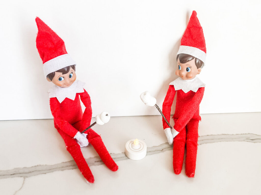 This image shows 2 elf on the shelf dolls pretending to be roasting mini marshmallows over an LED votive candle. This is one of the many elf on the shelf ideas for home included in this post.