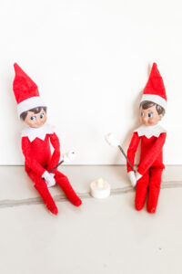This image shows 2 elf on the shelf dolls pretending to be roasting mini marshmallows over an LED votive candle. This is one of the many elf on the shelf ideas for home included in this post.