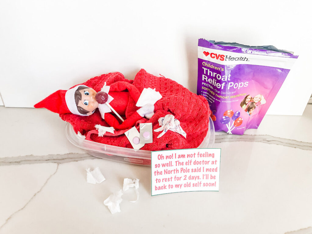 This image shows an elf on the shelf doll laying in a Tupperware container bed eating a throat relief pop, with medicine on him, and fake mini tissues surrounding him. The note says Oh No! I am not feeling so well. The elf doctor at the North Pole said I need to rest for 2 days. I’ll be back to my old elf self soon! This is one of the many elf on the shelf ideas for home included in this post.