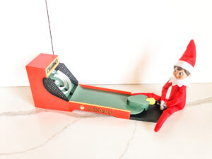 This image shows an elf on the shelf doll playing with a miniature size skeeball toy. This is one of the many elf on the shelf ideas for home included in this post.