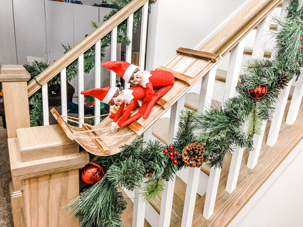 This image has 2 elves on a decorative wooden sled pretending to sled down the stairwell. This is one of the many elf on the shelf ideas for home included in this post.