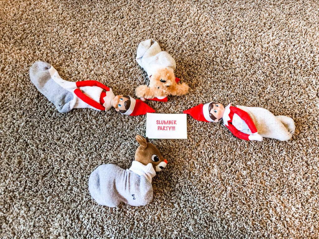 In this image, there are elf on the shelf dolls “sleeping” in socks with 2 stuffed animals also “sleeping” in socks. In the middle is one of the free printable notecards that says slumber party. This is one of the many elf on the shelf ideas for home included in this post.