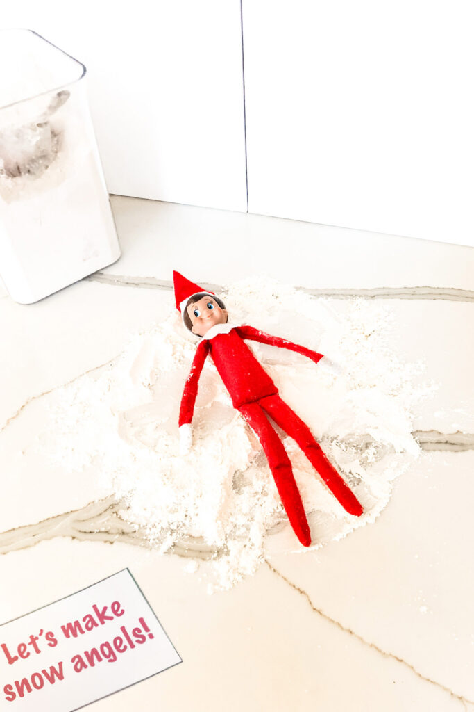 In this image, an elf on the shelf doll is making snow angels in flour. This is one of the many elf on the shelf ideas for home included in this post.