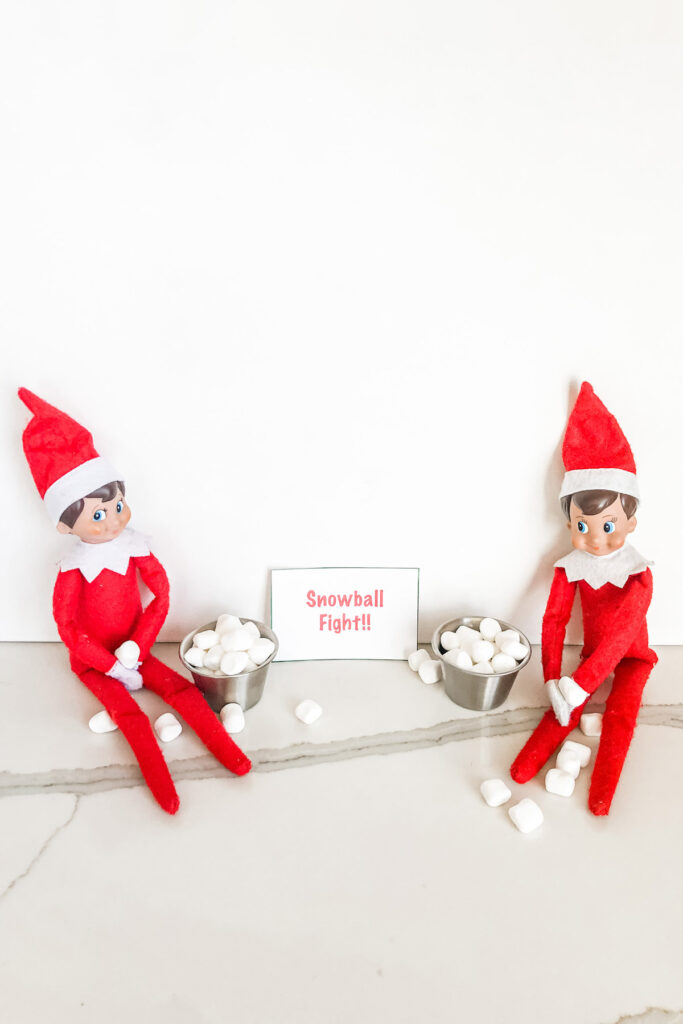 In this image, there are 2 elves with small metal containers of mini marshmallows. There is a sign that says snowball fight. This is one of the many elf on the shelf ideas for home included in this post.
