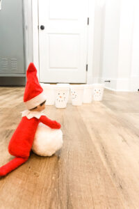 In this image you can see the back of an elf on the shelf with a white fake snowball and some white styrofoam cups with snowman faces on them pretending to bowl. This is one of the many elf on the shelf ideas for home included in this post.
