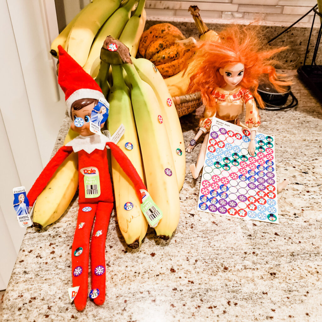 In this image, an elf on the shelf doll is sitting on the counter with an Anna doll and a sheet of stickers. Both dolls are covered in stickers. This is one of the many elf on the shelf ideas for home included in this post.