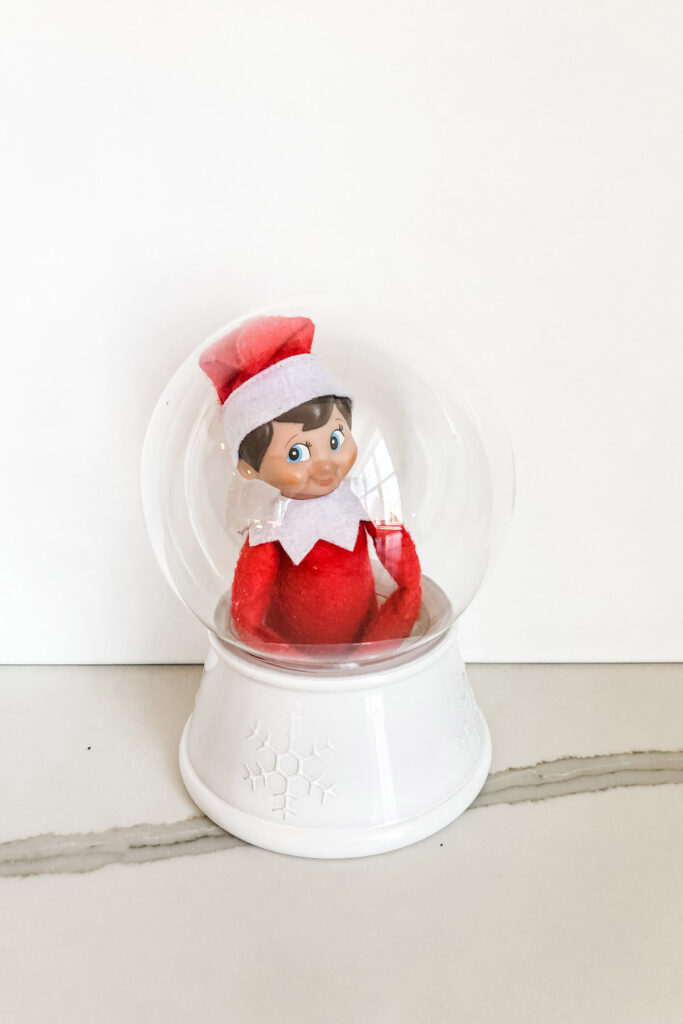 In this image, there is an elf on the shelf doll stuck inside of a snow globe. This is one of the many elf on the shelf ideas for home included in this post.