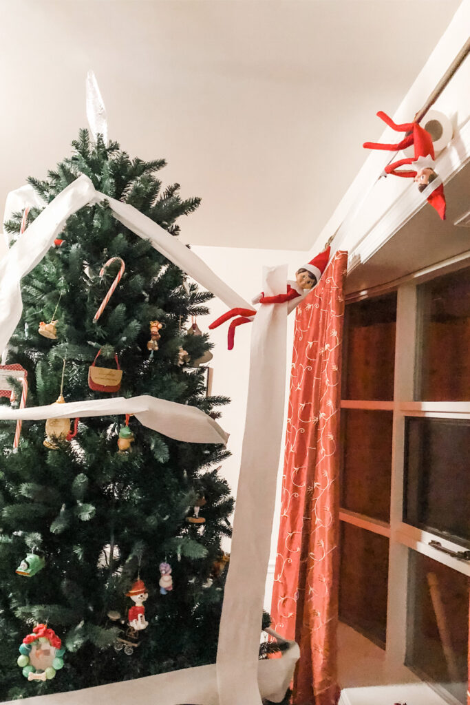 n this image, there are 2 elf on the shelf dolls that have wrapped toilet paper around a Christmas tree. One elf is hanging in the toilet paper like it’s a hammock. The other is hanging from a curtain rod upside down holding onto the paper. This is one of the many elf on the shelf ideas for home included in this post.