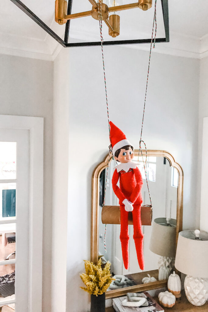 In this image, an elf on the shelf is sitting on an empty toilet paper tube that is hanging from a light fixture. This is one of the many elf on the shelf ideas for home included in this post.