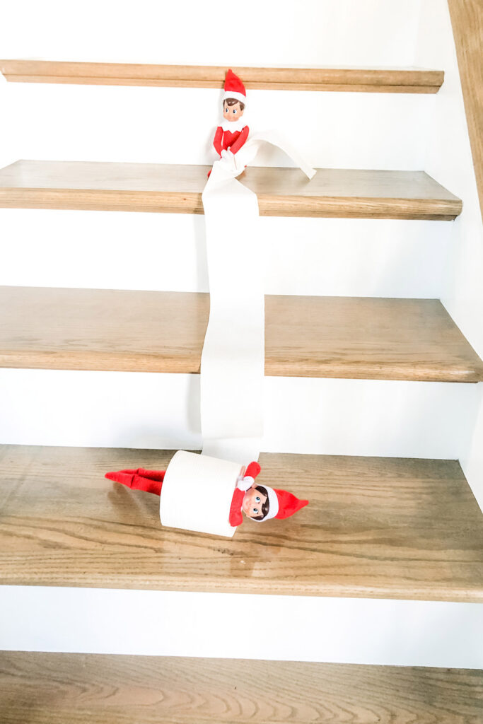 In this image, an elf on the shelf doll is rolling down the stairs stuck in a roll of toilet paper. A second elf is sitting higher up on the stairs holding the other end of the toilet paper. This is one of the many elf on the shelf ideas for home included in this post.