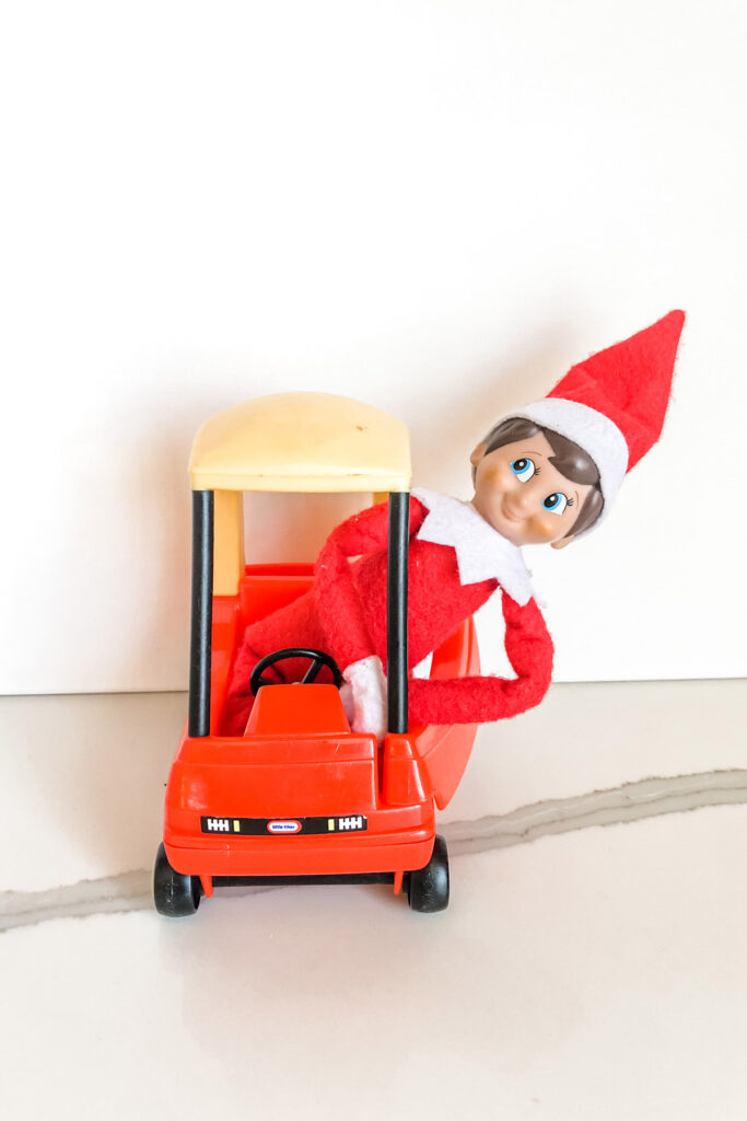 In this image, an elf on the shelf doll is sitting inside of a mini Little Tikes red car. This is one of the many elf on the shelf ideas for home included in this post.