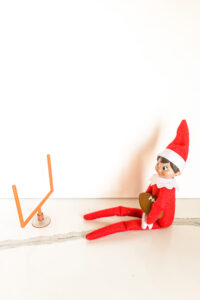 In this image, an elf on the shelf doll is holding a mini toy football and sitting in front of a pretend mini football goal. This is one of the many elf on the shelf ideas for home included in this post.